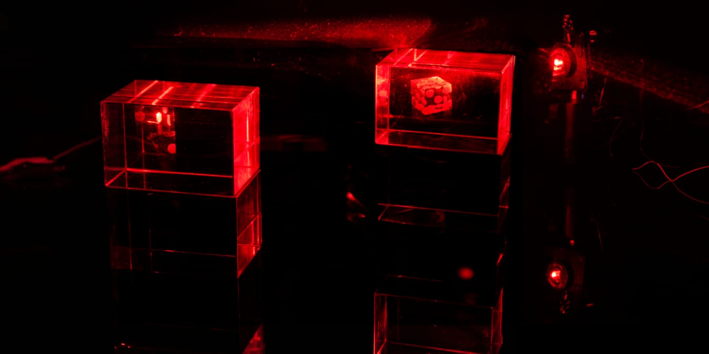 Two glowing holograms are lit by red lasers. One image of a single die is rendered in full while the second image of several dice is displayed in the same light path and partially occluded by the shadow of the first mount.
