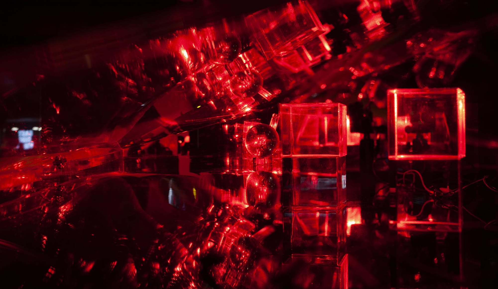 Clear cubes and a crystal ball lit by red lasers on black ground reflect into an endless circular pattern in a long clear sloped case that recedes into space.