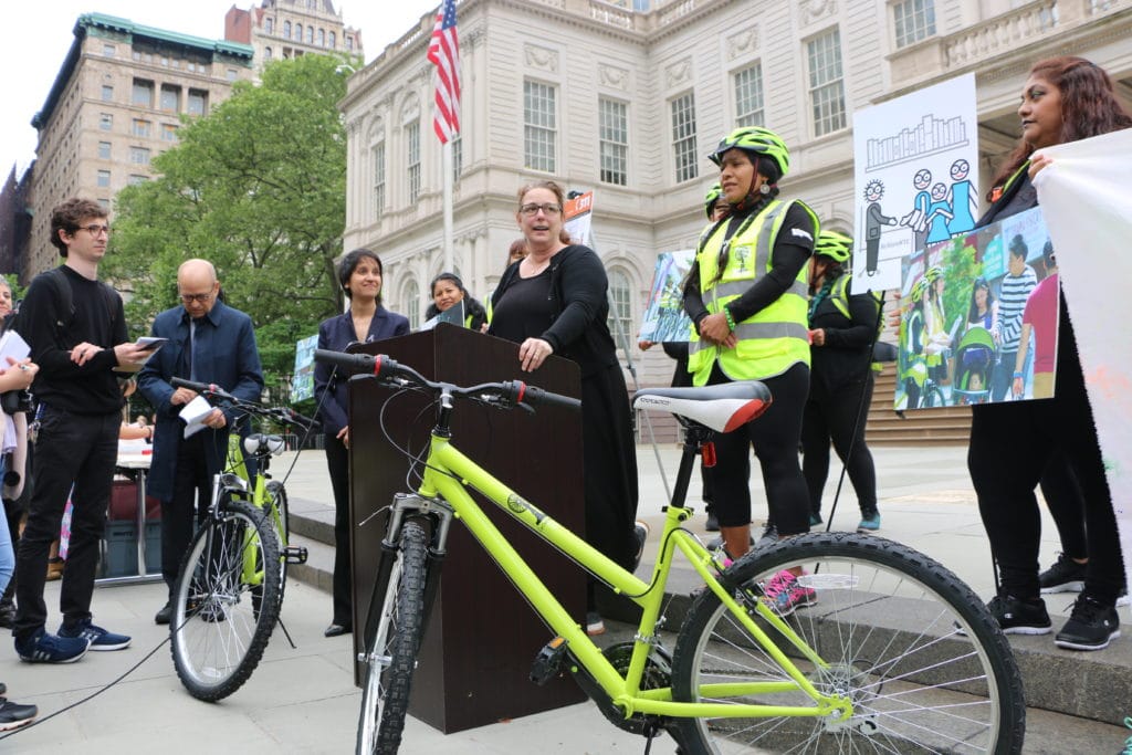 Tania Bruguera stands at a podium with administrators and cyclists. a lime green bicycle sits, riderless, in the foreground.