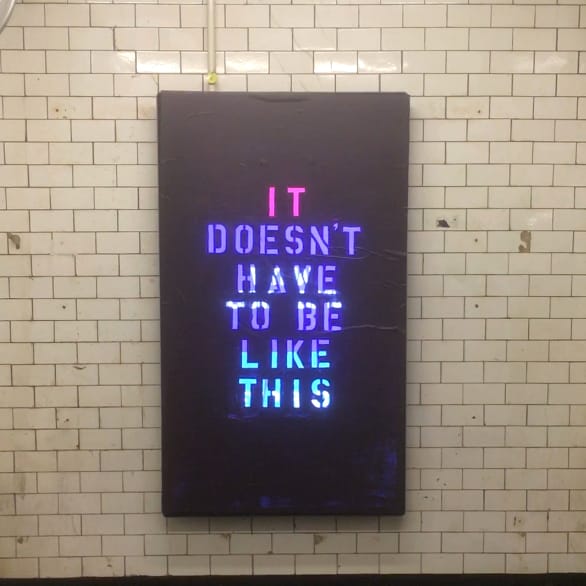 An LSD screen is covered by a black stencil covering featuring the words "It doesn't have to be like this" in pink, purple, and blue light, set against a dirty tiled wall underground in the subway.