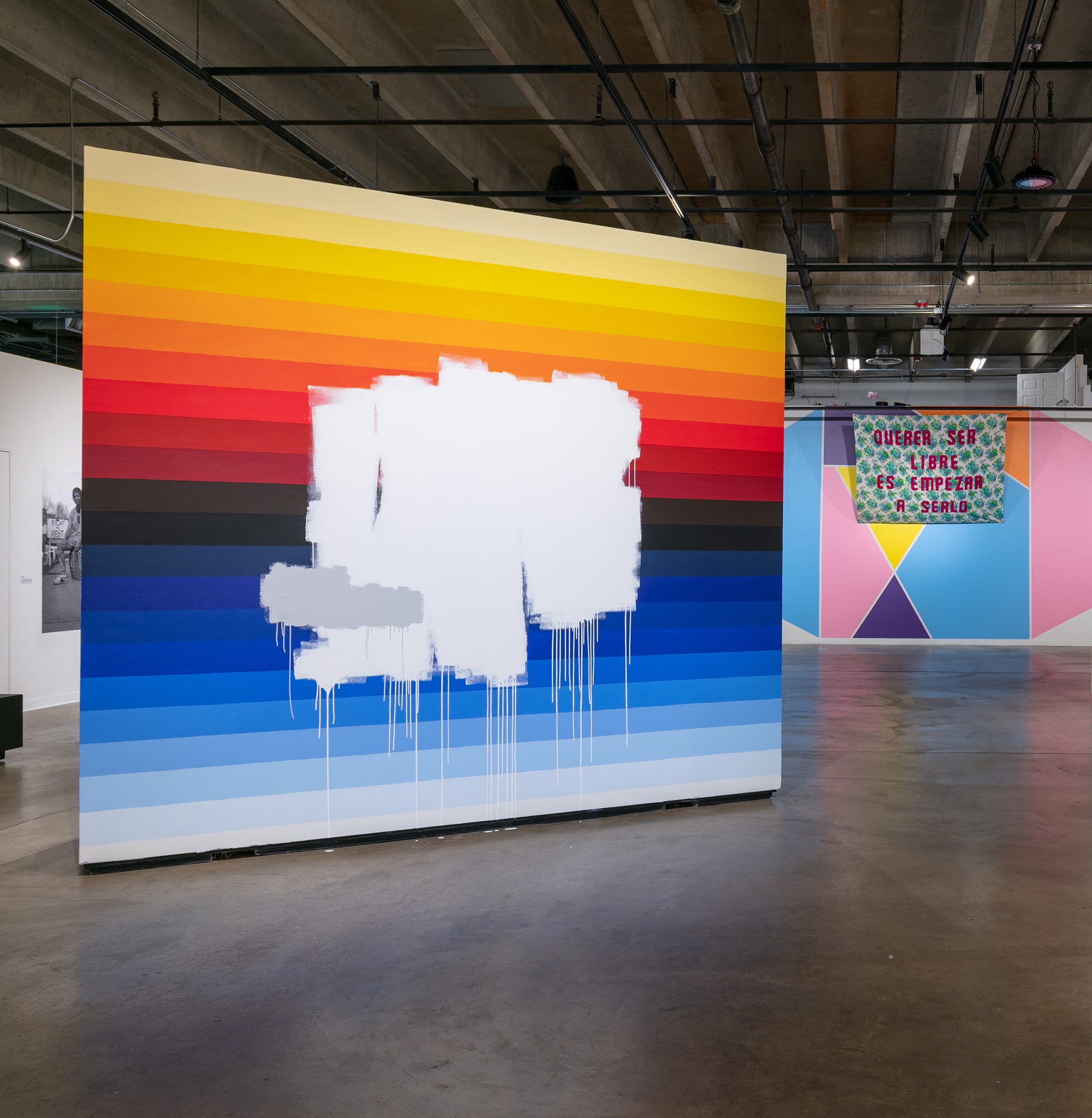 View onto a large gallery space with concrete floors. In the foreground, a monumental freestanding streetart-inspired work features horizontal gradient stripes that go from shades of yellow, orange, red, brown, black, and blue. In the middle of the canvas, messy rolls of white paint drip over. In the background, to the right, hangs a green and white-patterned textile with "QUERER LIBRE ES EMPREZAR A SERLO" in red stitched over. Behind the textile is a geometric-style painted background of blue, purple, yellow, orange, and pink.
