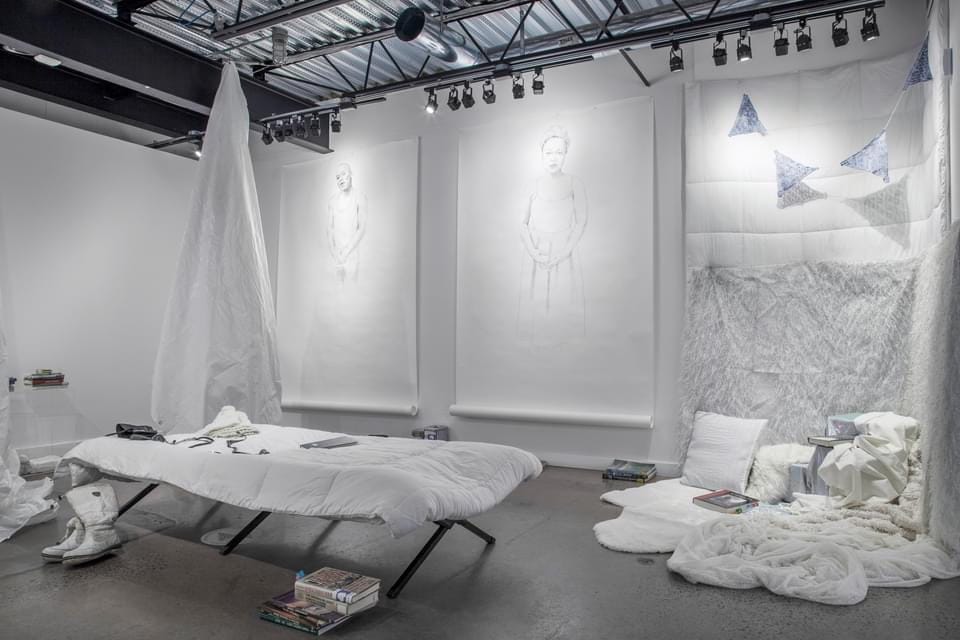 View onto a white-walled gallery space with concrete flooring. Below a bar of track lighting, hang two large-scale, seemingly unfinished drawings of figures. Next to the two paper works is a white comforter, which touches down to the floor and is augmented by white pillows and white bed sheets. At the foreground, a cot with white bedding stands, with a pair of white boots. Scattered throughout the space are small stacks of books.