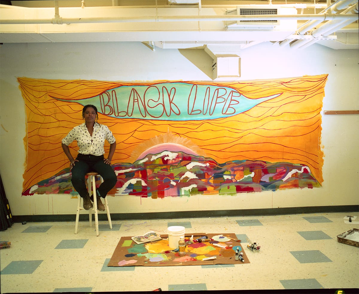 View onto a classroom-like space. At the foreground, on the floor, rests paint materials. Behind, sitting on a stool, is the artist, a femme-presenting Black person. Behind this person hangs a large, colourful artwork. There is an orange sky with red lines, above an abstract, multi-coloured city skyline with the sun peaking above. At the top, In red outlined text behind a blue background reads "BLACK LIFE."
