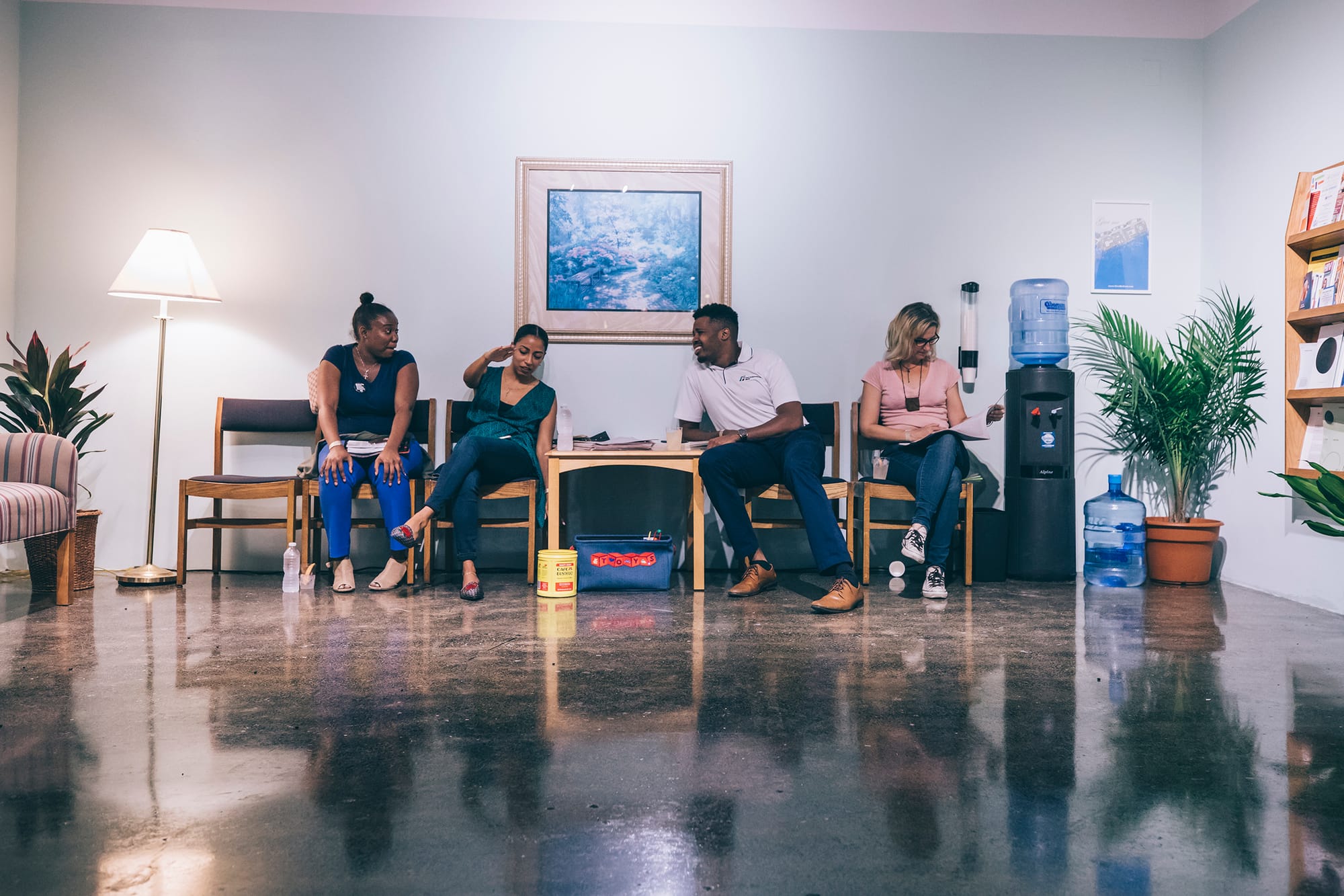 View onto an institutional looking room with chairs in a row separated by a wooden table covered with reading materials. Four people sit against a light-green wall. The three on the left are talking. One is a masculine-presenting Black person and the other two are femme-presenting Black people. A femme-presenting white person is reading a newspaper on the right. In the room’s left corner is a lamp and a potted large-leafed plant. In the room’s right corner is a water cooler and a similar plant next to a wall-mounted brown bookshelf full of publications.