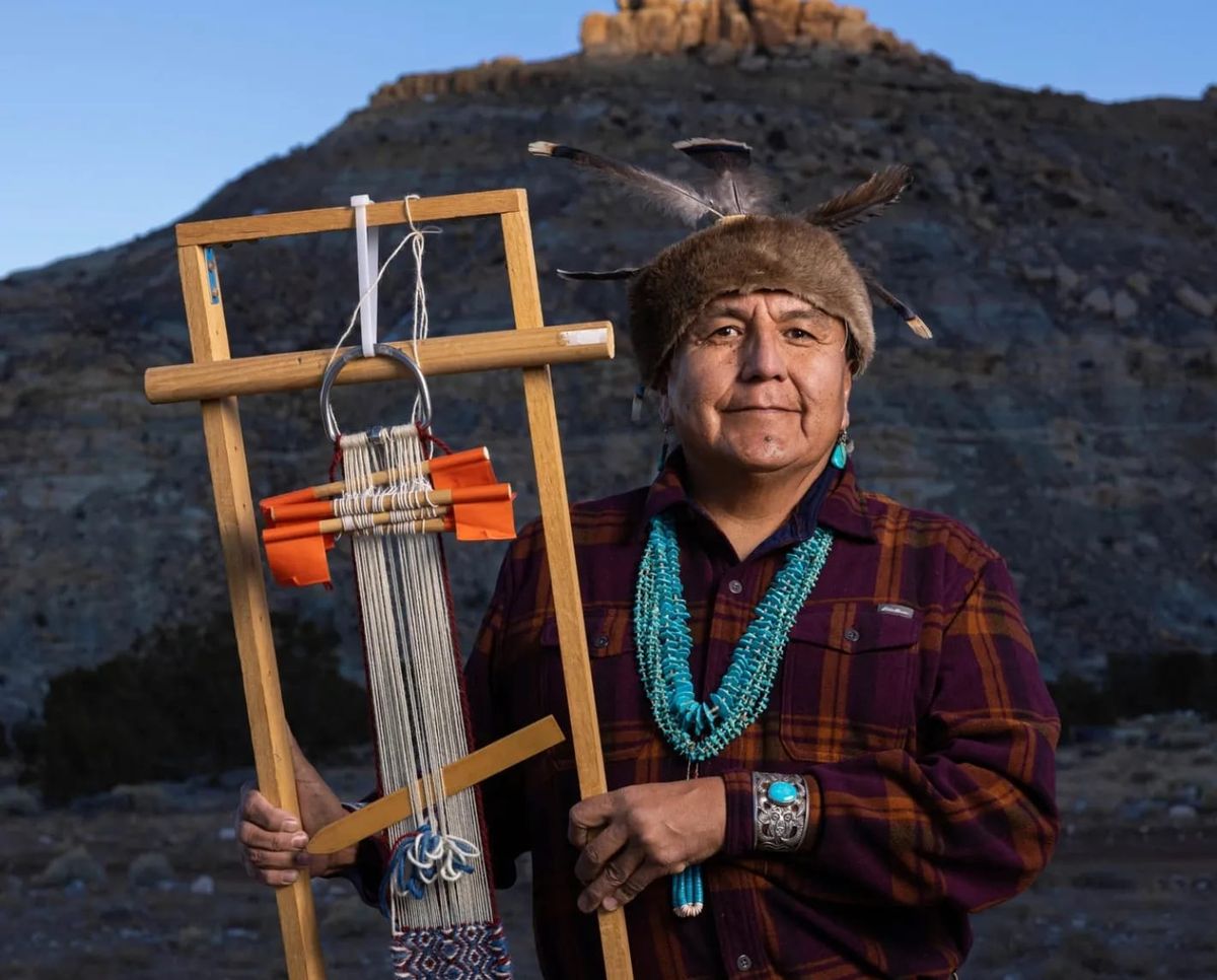 Portrait of Roy Kady in turquoise and a feathered cap, holding a weaving loom before a desert mesa