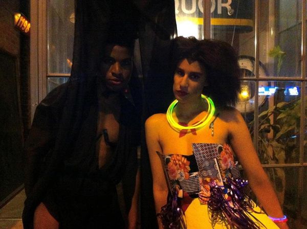 Lavish Bat, in black veil, stands next to reeraw, wearing an angular wig and colourful paper dress accessorized with a glowst