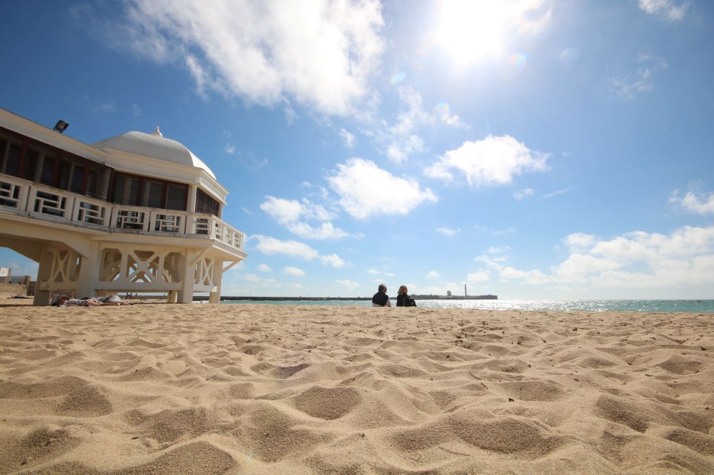 A couple sitting on a golden sand beach at La Caleta in Cadiz, with a white pier - best beaches in Spain