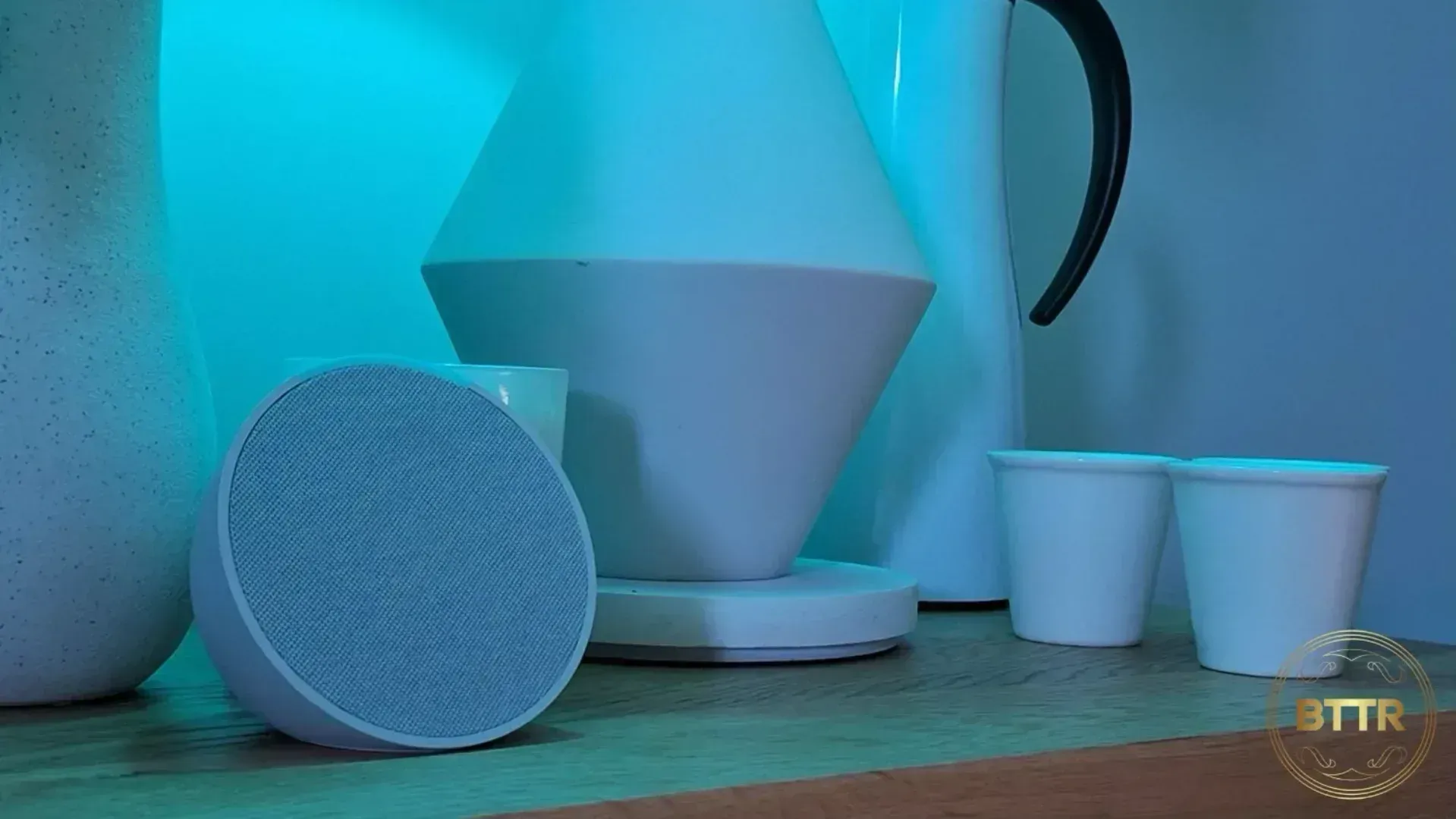 The Amazon Echo Pop on a bedside table next to a lamp and some cups