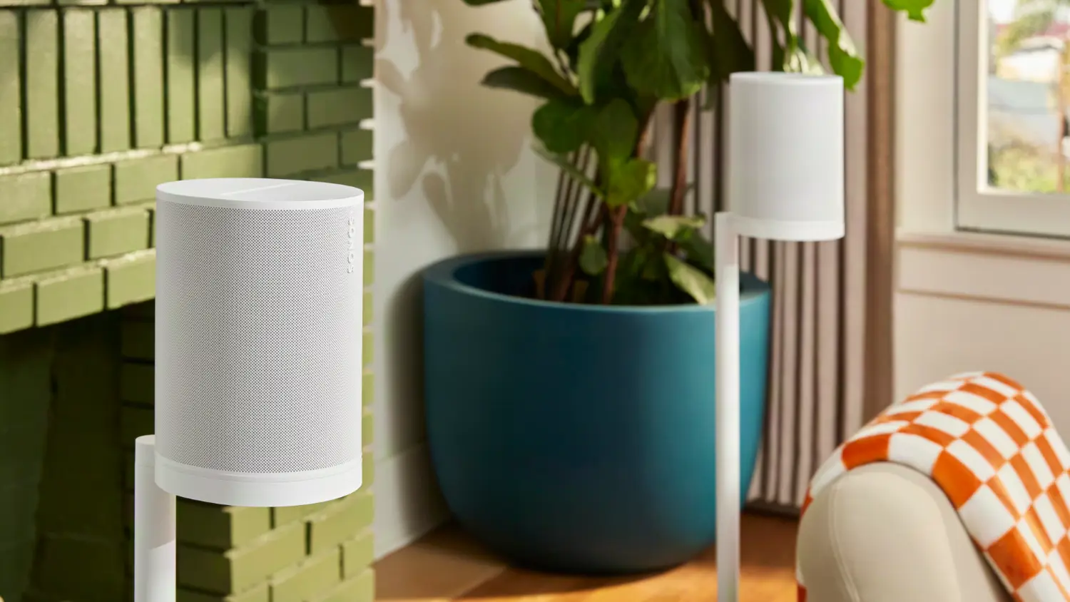 Two Sonos Era 100 SL speakers on stands behind a couch, with a plant and a fireplace in the background