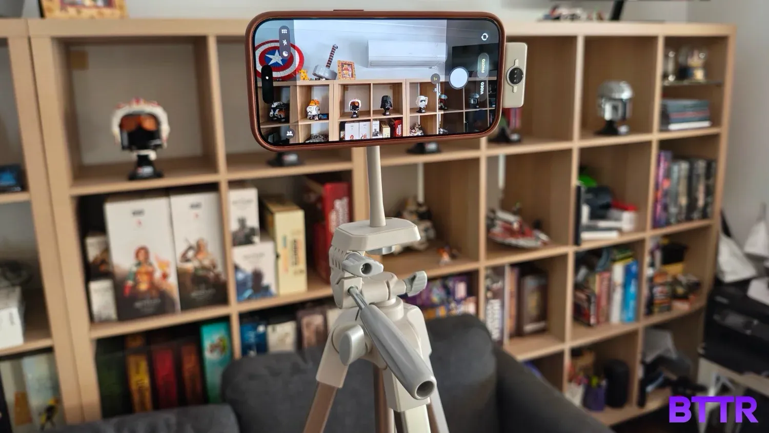 An iPhone 17 attached to the magentic camera grip of teh Belkin Stage Creator Kit's tripod in front of a bookshelf full of Lego Star Wars helmets and board games.