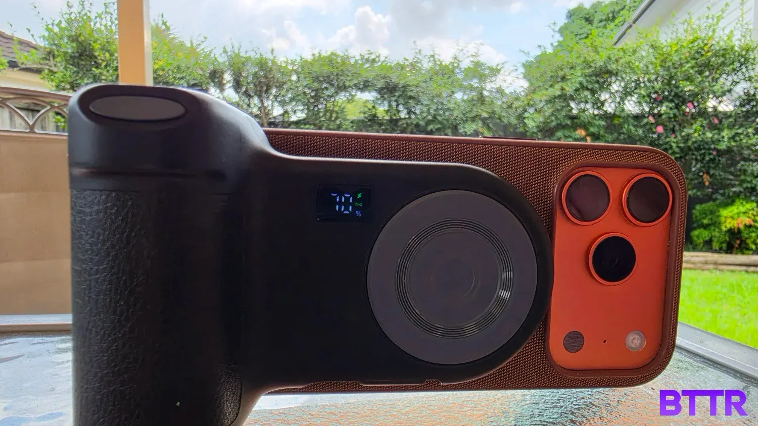 The Belkin Stage PowerGrip on a table with a garden in the background, connected to the iPhoen 17 Pro