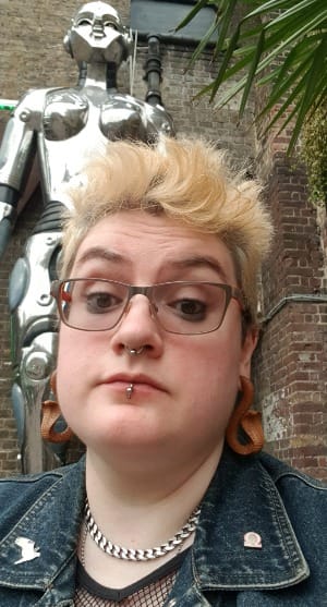 Me (G. B. Bard) looking a little frazzled beneath a towering chrome femmebot outside Cyberdog in London. I have fluffy bleached hair, wooden snake earrings and a denim vest on.