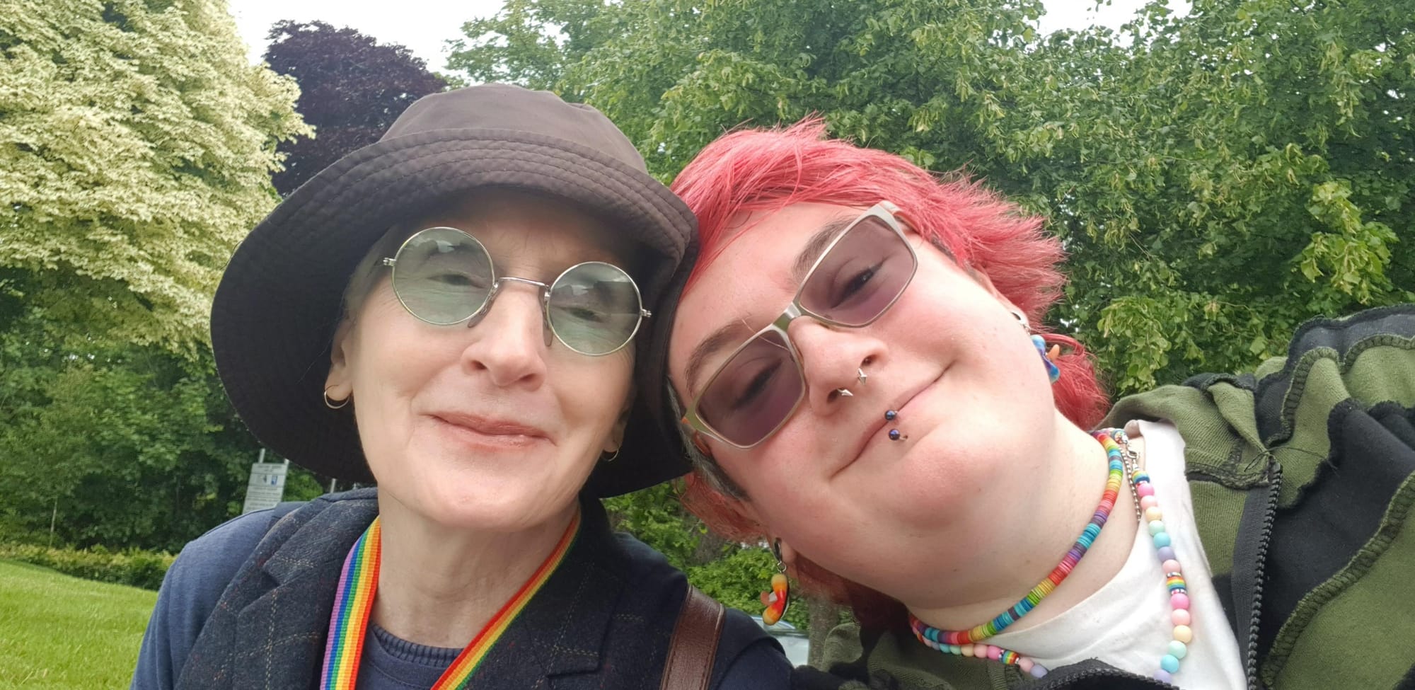 Two happy people leaning their heads together with trees & grass behind. My mum is on the left in round green sunglasses & khaki sun hat, I’m on the right with pink-red hair, rainbow chokers & green/black striped hoodie.