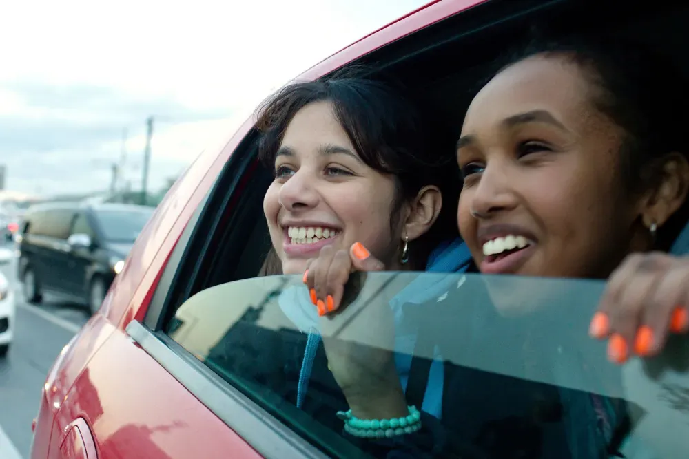 A British Pakistani girl and a Black British girl with neon orange nail polish grin excitedly as they lean out the window of a red car on a highway. They seem thrilled to be travelling.