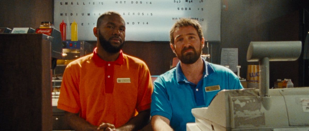 A black man in an orange retail t-shirt looks sceptically at the camera, his nametag reads Lorenzo. Beside him a white guy with a beard, called Hunter, looks thoroughly fed up. They are behind a cinema concession counter.