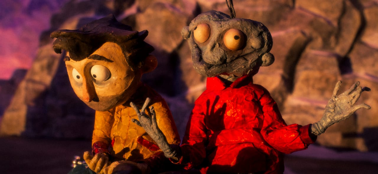 Modelled rocky scenery surrounds stop-motion of a tired-looking man with messy brown hair and stubble in a yellow jacket. He is sitting beside a ghoulishly smiling, blueish rotting corpse in a bright red padded jacket. The sunrise is falling across their faces.
