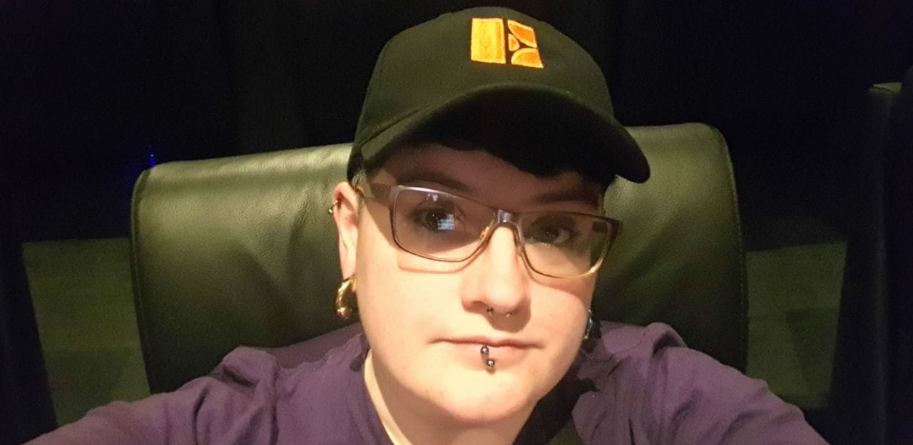 G.B.Bard smiling sagely from a black leatherette recliner chair, wearing silvery glasses, chunky metallic earrings, facial jewellery and sporting a black cotton baseball cap with an embroidered 'E' on in orange. I am also wearing a lovely purple t-shirt.