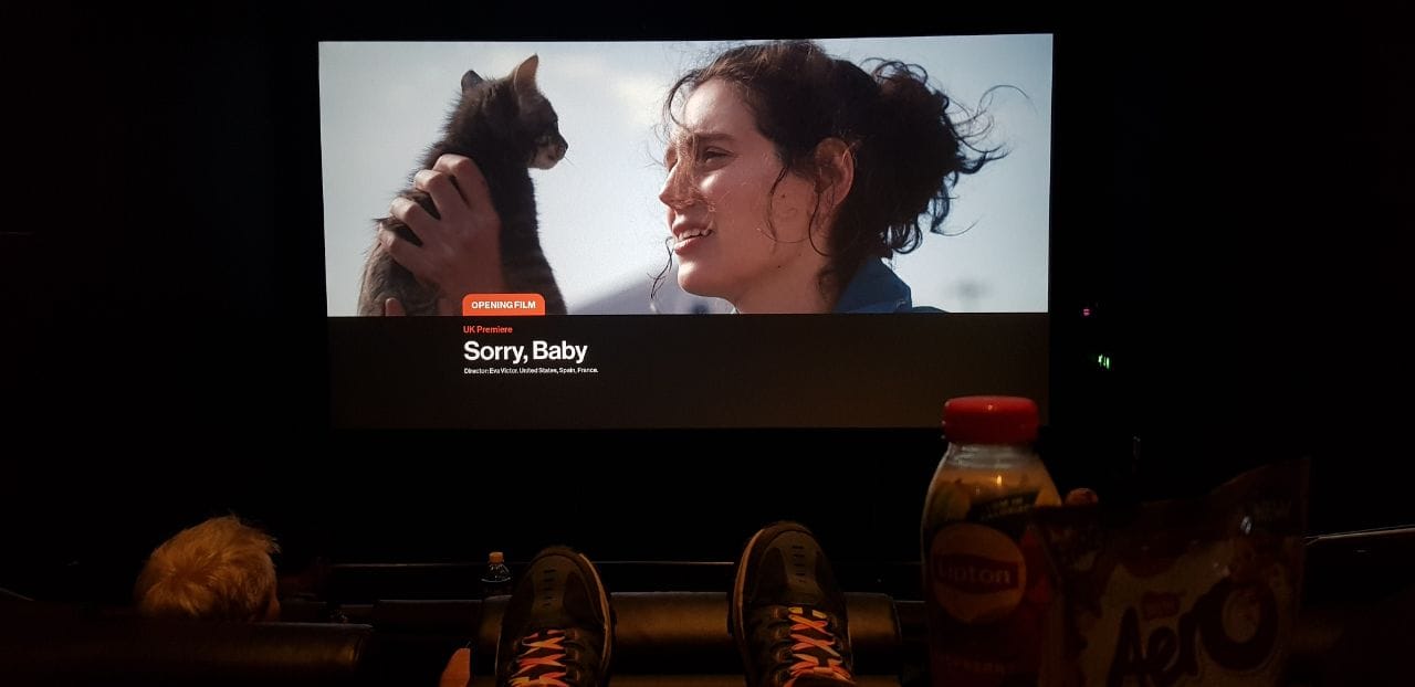 An image taken in a darkened cinema with black leatherette recliner seating. You can see a bottle of Lipton Iced Tea and some Aero Melts on the armrest tray, and the toes of my blue Sketchers with rainbow laces stretched out in front. On the screen is a still for the Opening Film "Sorry, Baby" with the female lead holding a smol tabby kitten up.