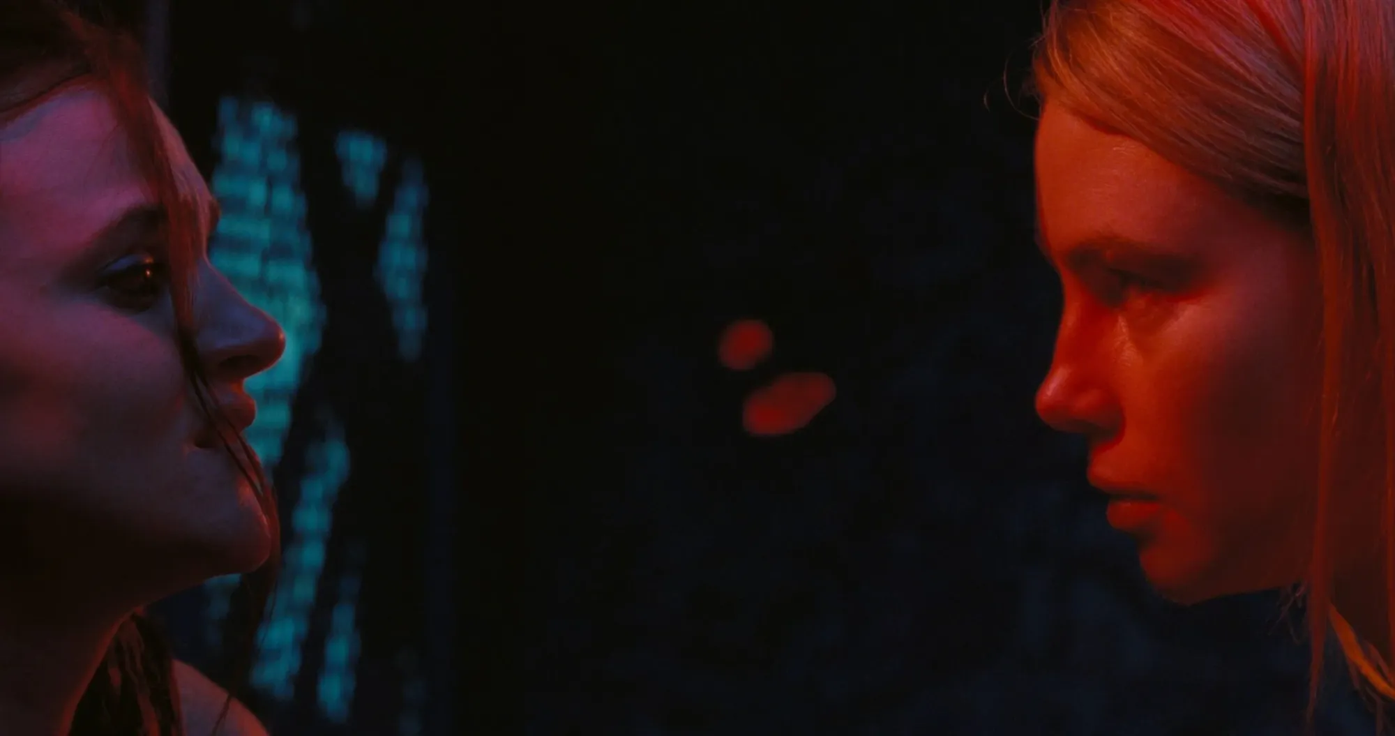 Lillian, a woman with dark hair and a witchy aesthetic looks worriedly across a near-black divide of space between her and the intense stare of Rose, a blond-haired girl cast in a red glow.