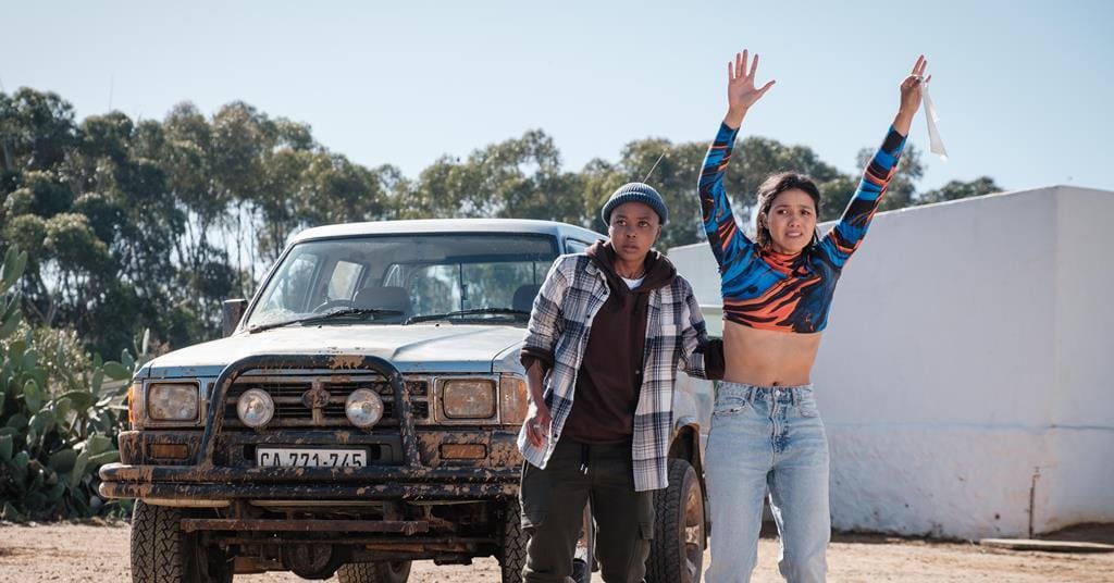 A non-binary black person in hoody and beanie hat stands hesitantly beside a mixed-race woman in neon dance crop-top waving a wet wipe as a white flag. They are standing in front of their Hilux in the South African sun.