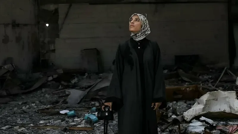 A young, muslim woman stands in a black cloak, with her glasses propped up on her black and white hijab, looking skyward. She clutches a serious photographic camera in one hand and stands amidst the rubble of a bombed-out building.