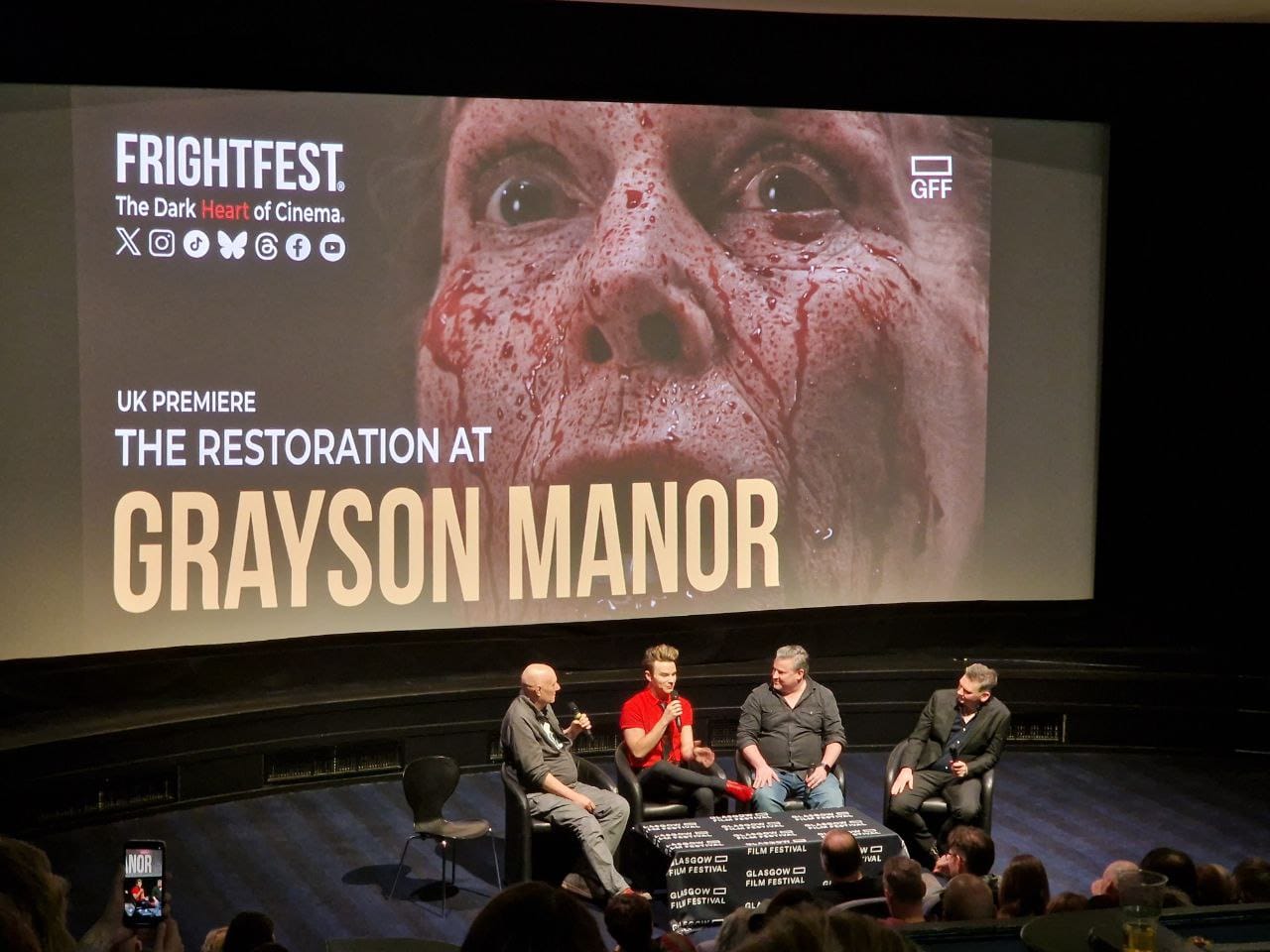 Four cast members and an interviewer sit in chairs by a film festival table at the foot of the GFT1 screen. On the screen is a blood-drenched woman in shock with the words UK Premiere: The Restoration at Grayson Manor.