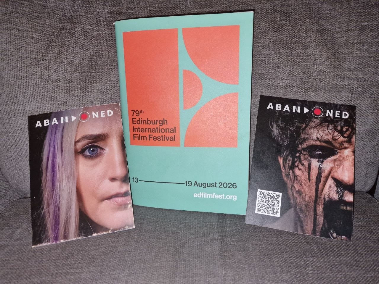 Orange & black EIFF logo on a turquoise background states "79th Edinburgh International Film Festival, 13-19 August 2026, edfilmfest.org". Either side of the programme are promo postcards from Abandoned. One has pre-scared Jess face on, the other has bile-drenched Adam face and a QR code.