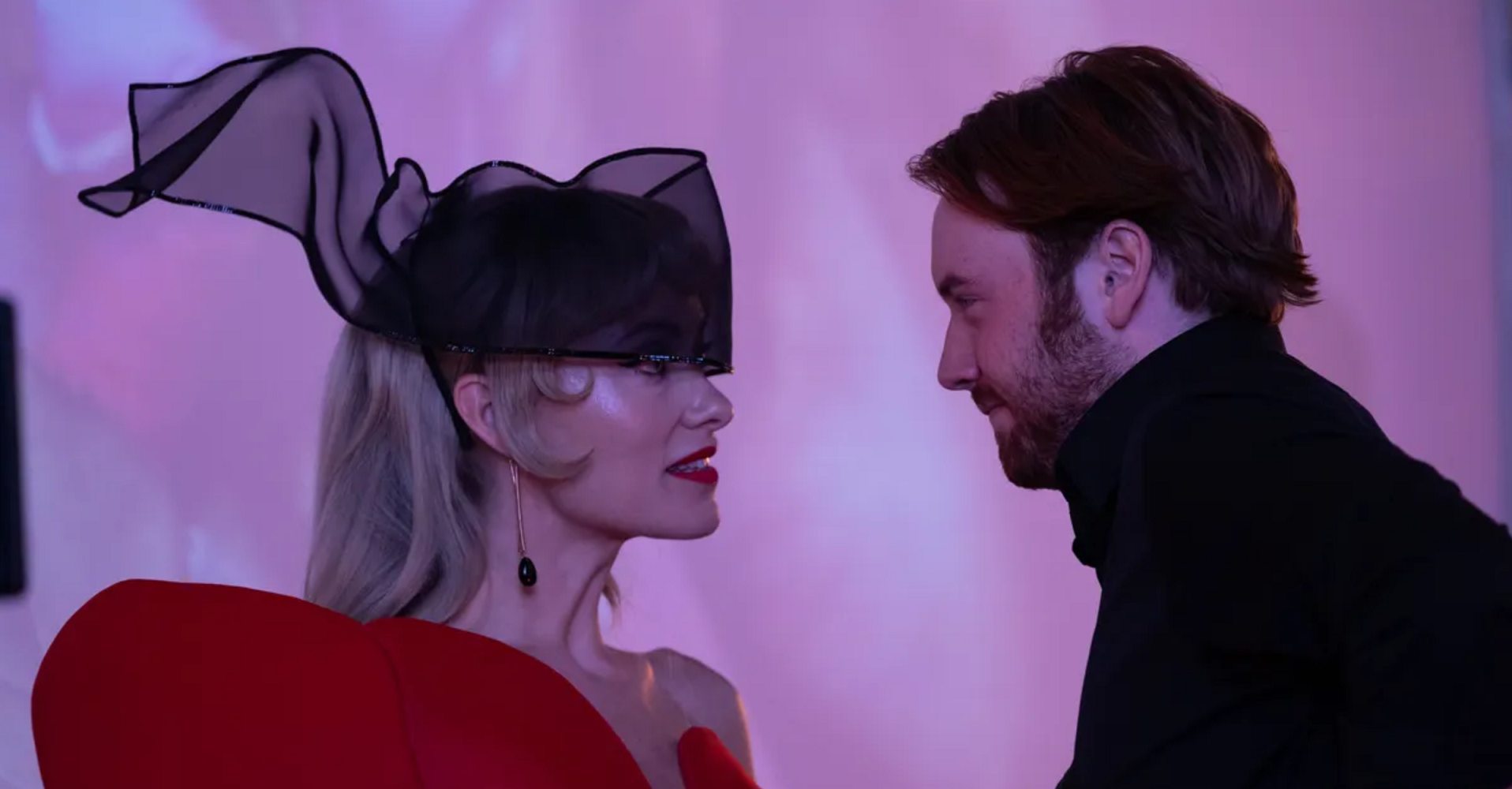 A high-class female artiste with blond hair, crystal drop earrings, red lipstick, an Avant Garde black ribbon hat and an asymmetric red dress looks intensely at a young man with ginger hair and beard in a black high-necked suit. Erotic pink flowers are being projected on the wall behind them.