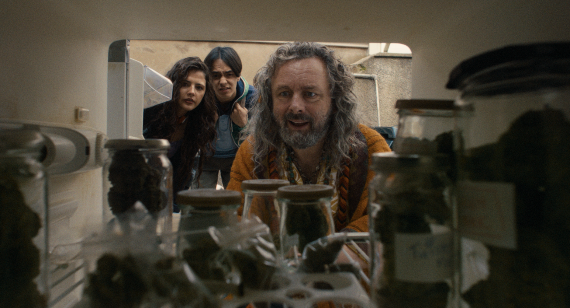 Picture taken from within a fridge filled with jars of magic mushrooms. Two teens, Maz and Elis peer curiously in from outside, whilst the very hippy store owner Shifty Gruff opens the fridge to inspect the wares.