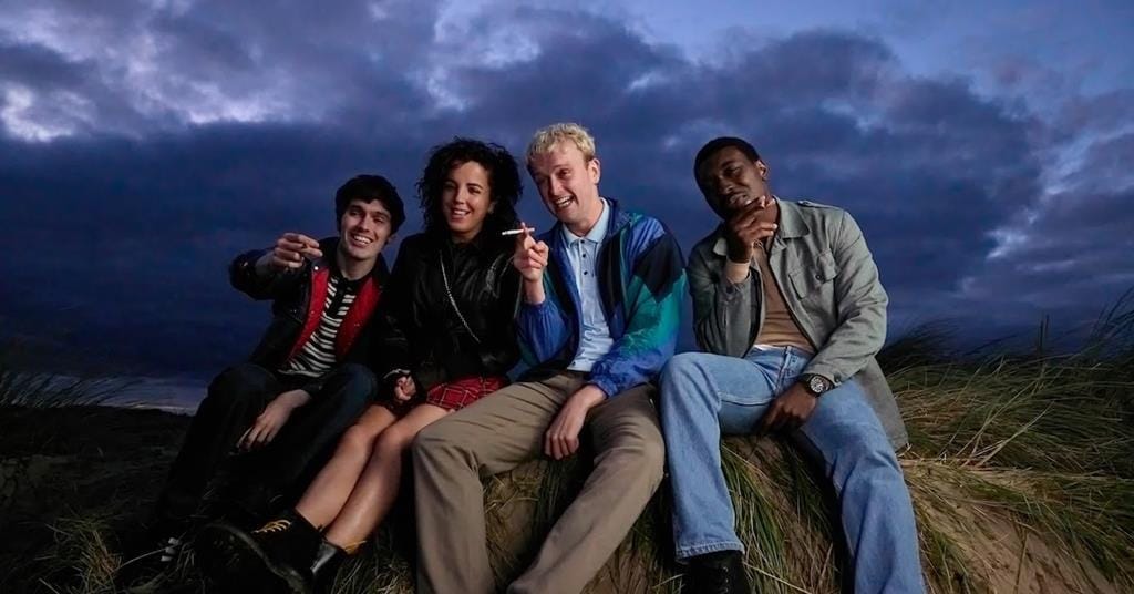 Four young adults in casual 90s rave garb sit gesturing, smoking cigs and giggling shoulder-to-shoulder on a grassy sandbank as dawn creeps across a cloudy sky.