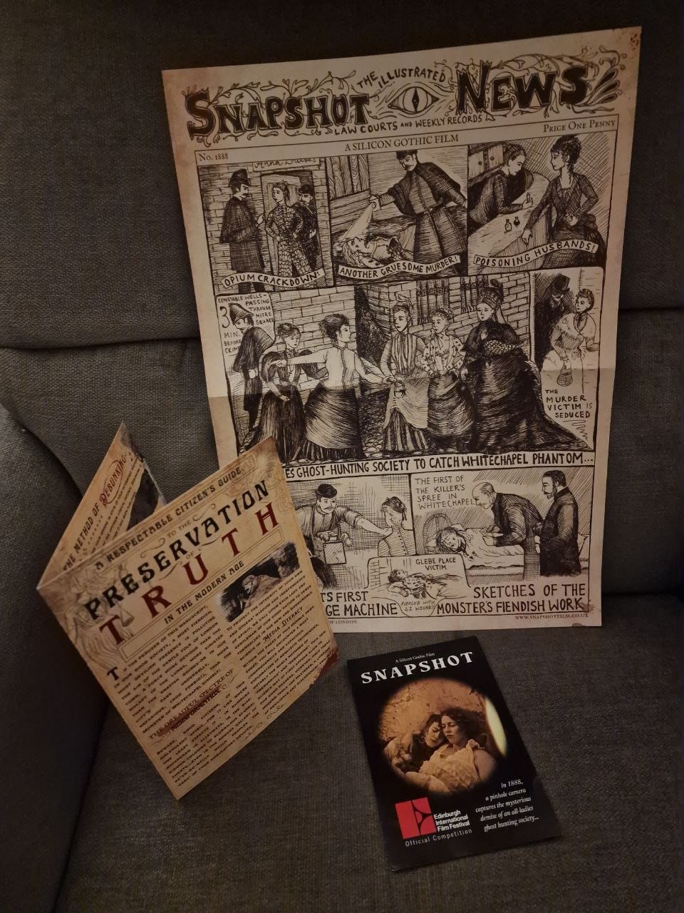 Promo materials placed on a grey fabric chair: central is an illustrated broadsheet in the 1888 style sensationalising the Ripper murders, to the left is a pamphlet titled "A Respectable Citizen's Guide To The Preservation Of Truth In The Modern Age" which describes methods of debunking disinformation, and at the base is a postcard poster of the film with pinhole sepia picture of two Victorian ladies embracing.