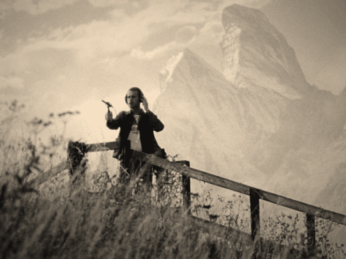 A foley artist with shaved hair and casual double-denim holds a mic up to nature and listens on his headphones by a fence overlooking a field of grass. In the background looms a mountain shaped like a crab's pincer. It is a beautiful day.