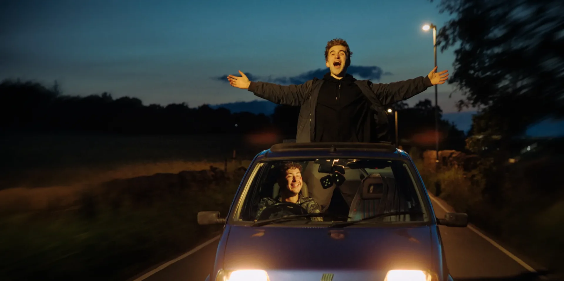A tiny blue car drives through the Yorkshire countryside with its lights on at twilight. One young lad grins as he drives the car with dice on the mirror, his mate stands through the skylight with his arms out & wind in his hair, yelling excitedly.