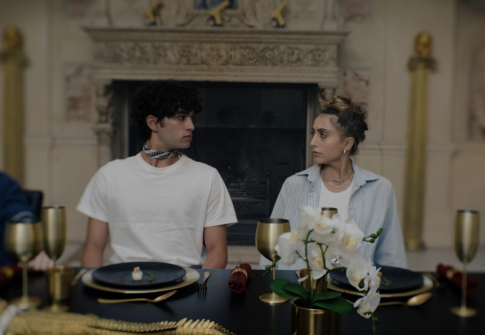 A young Mediterranean man with curly black hair, a neckerchief and white t-shirt stares curiously at a beautiful non-binary femme with gold necklaces and a stripy shirt sitting at the dining table with him. The room they are in is a grand old estate with an ornate fireplace.