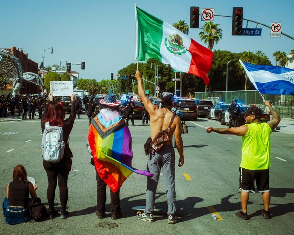 5 protesters stand at an L.A. intersection facing a wall of police. A Trans-inclusive Pride flag, Mexican flag & El Salvador flag wave proudly. A sign reads "Remember Your Oath".