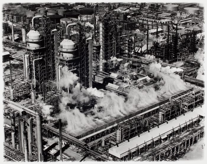 High-angle black-and-white photograph of a massive industrial refinery. Large vertical cracking towers and cooling units are enmeshed in a dense network of pipes and scaffolding, with billowing steam obscuring parts of the machinery, emphasizing the intense complexity of oil processing.