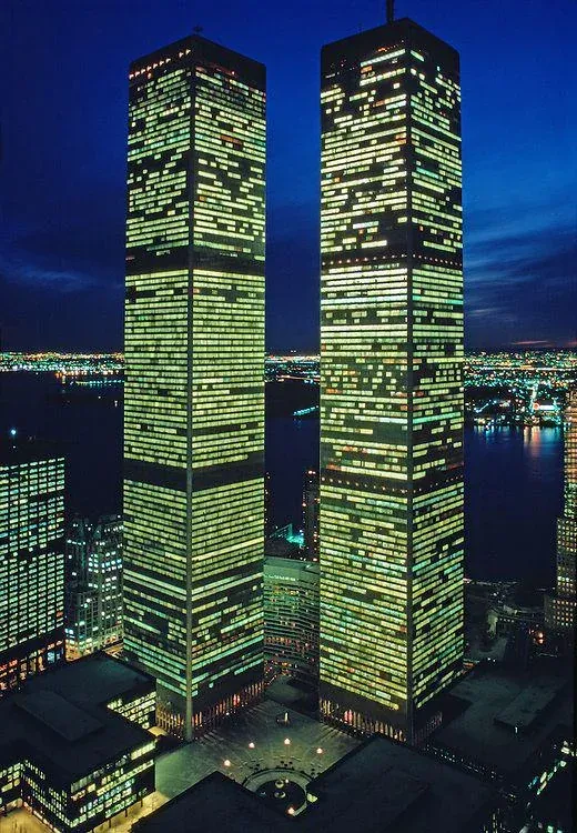 A nighttime photograph of the Twin Towers with many of the office windows glowing with yellow light. The surrounding city lights and the dark water of the harbor are visible in the background.