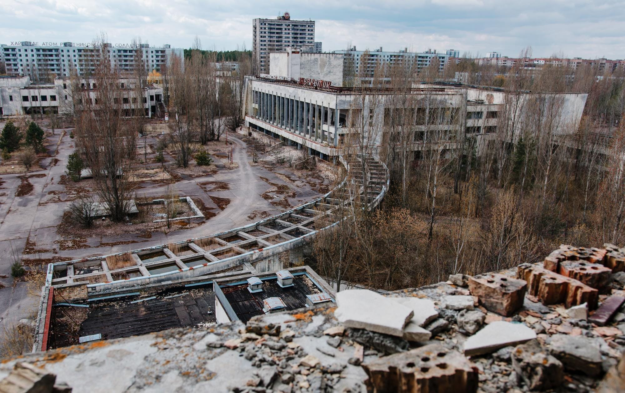Elevated vantage over an abandoned urban plaza, showing a decaying concrete cultural center with a ruined, skeletal walkway surrounded by barren trees and desolate apartment blocks.