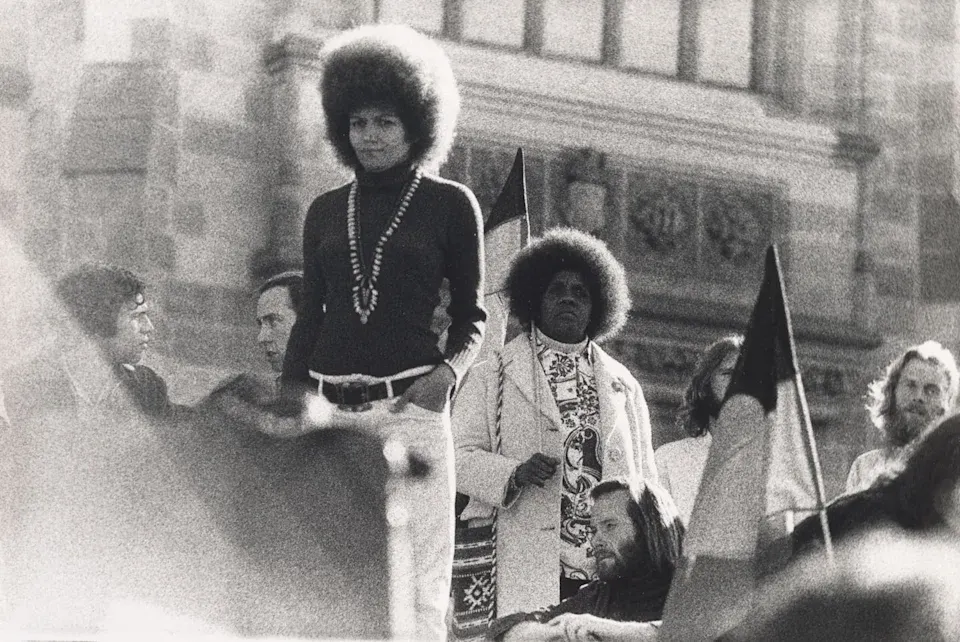 Bobbi Sykes speaks forcefully from a podium, her arms resting defiantly at her waist while she gazes off-camera, flanked by flags in a confrontation with the systemic violence of the colonial administration.