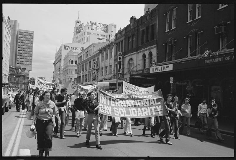 A large crowd marches down a city street holding a banner for the International Gay Solidarity Group.