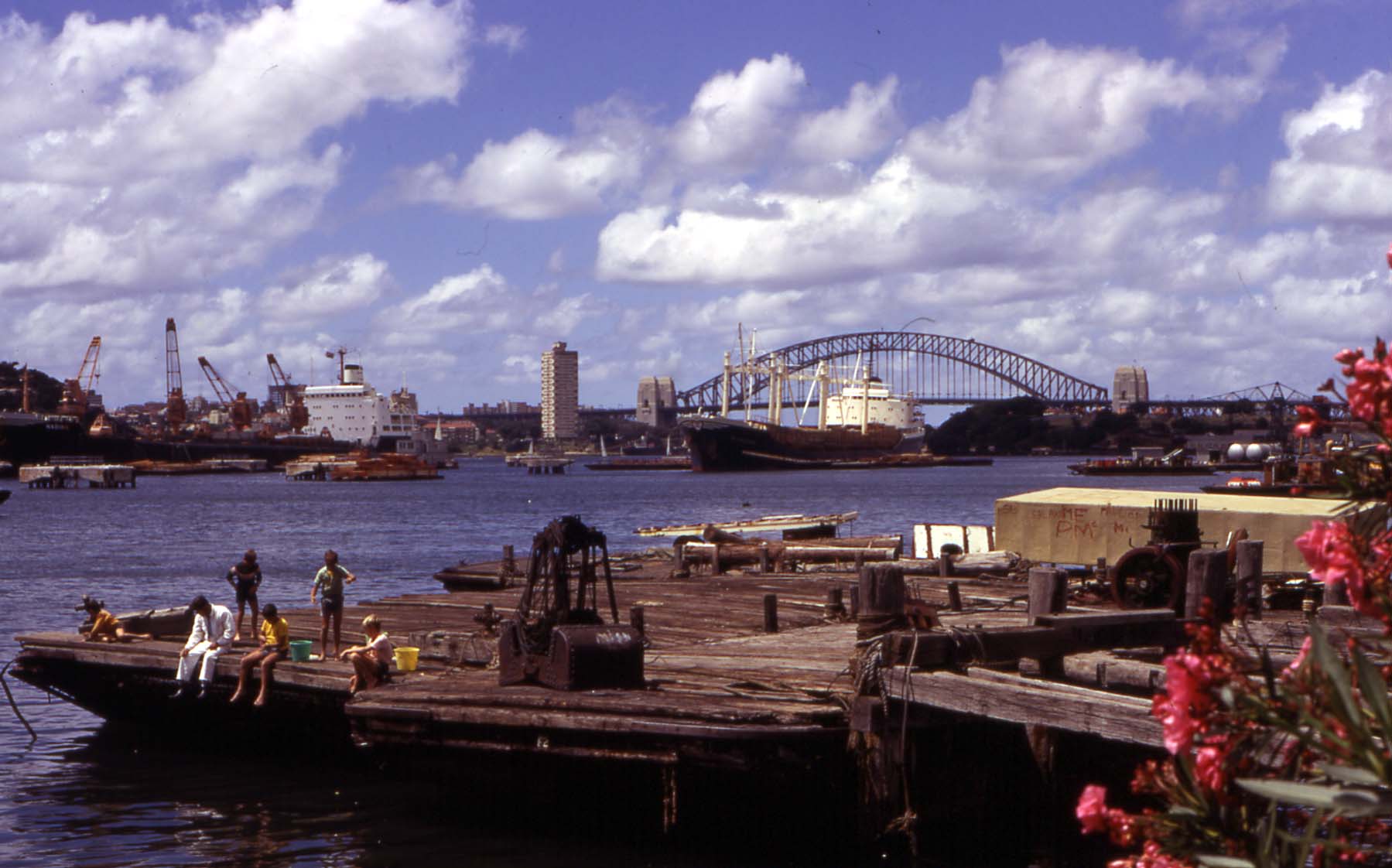 Children sitting on a wooden pier looking out at industrial shipping vessels and the Sydney Harbour Bridge.