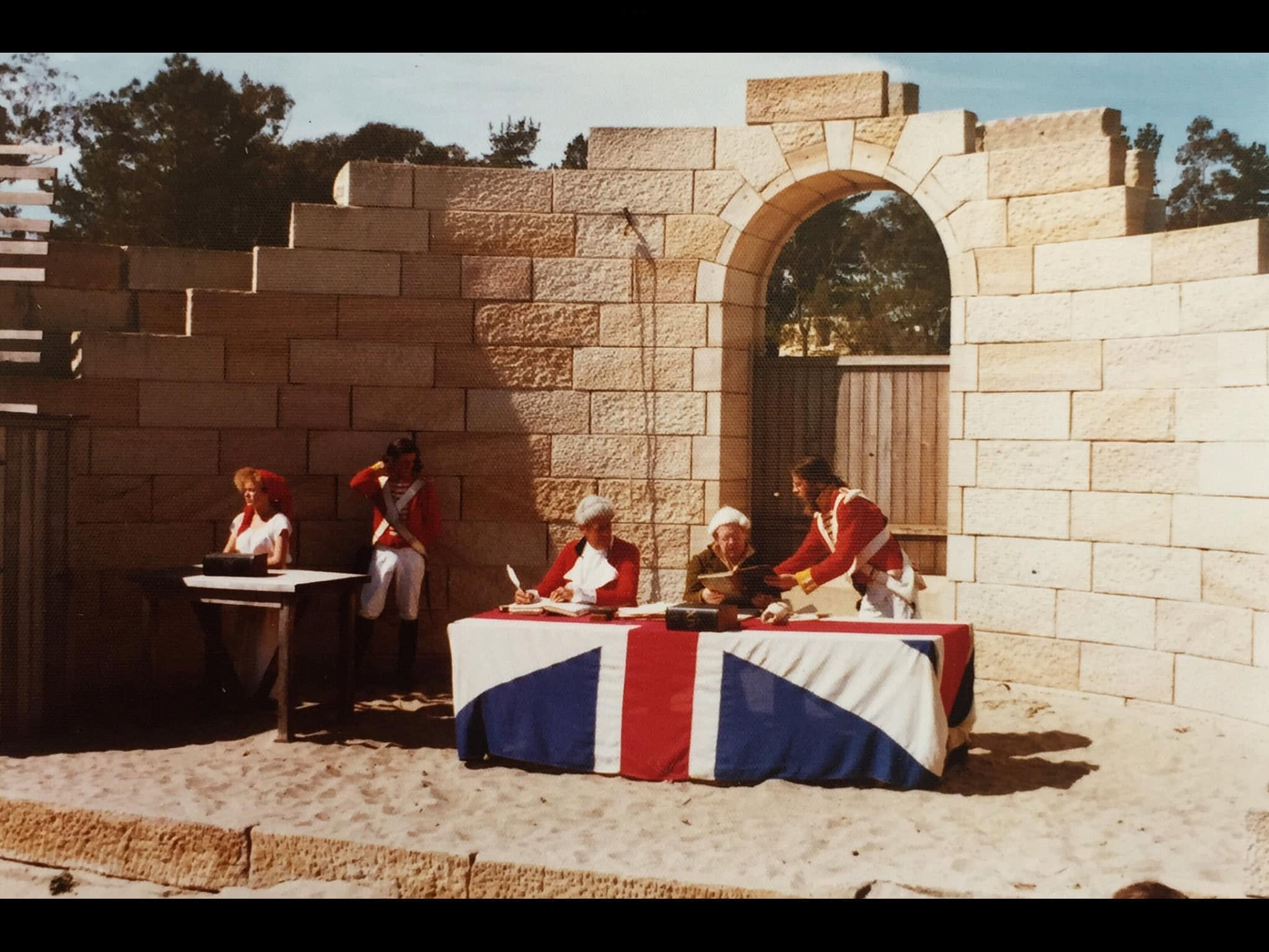 Actors in colonial British military uniforms staging a historical recreation behind a Union Jack flag, sanitizing imperial history for consumption.