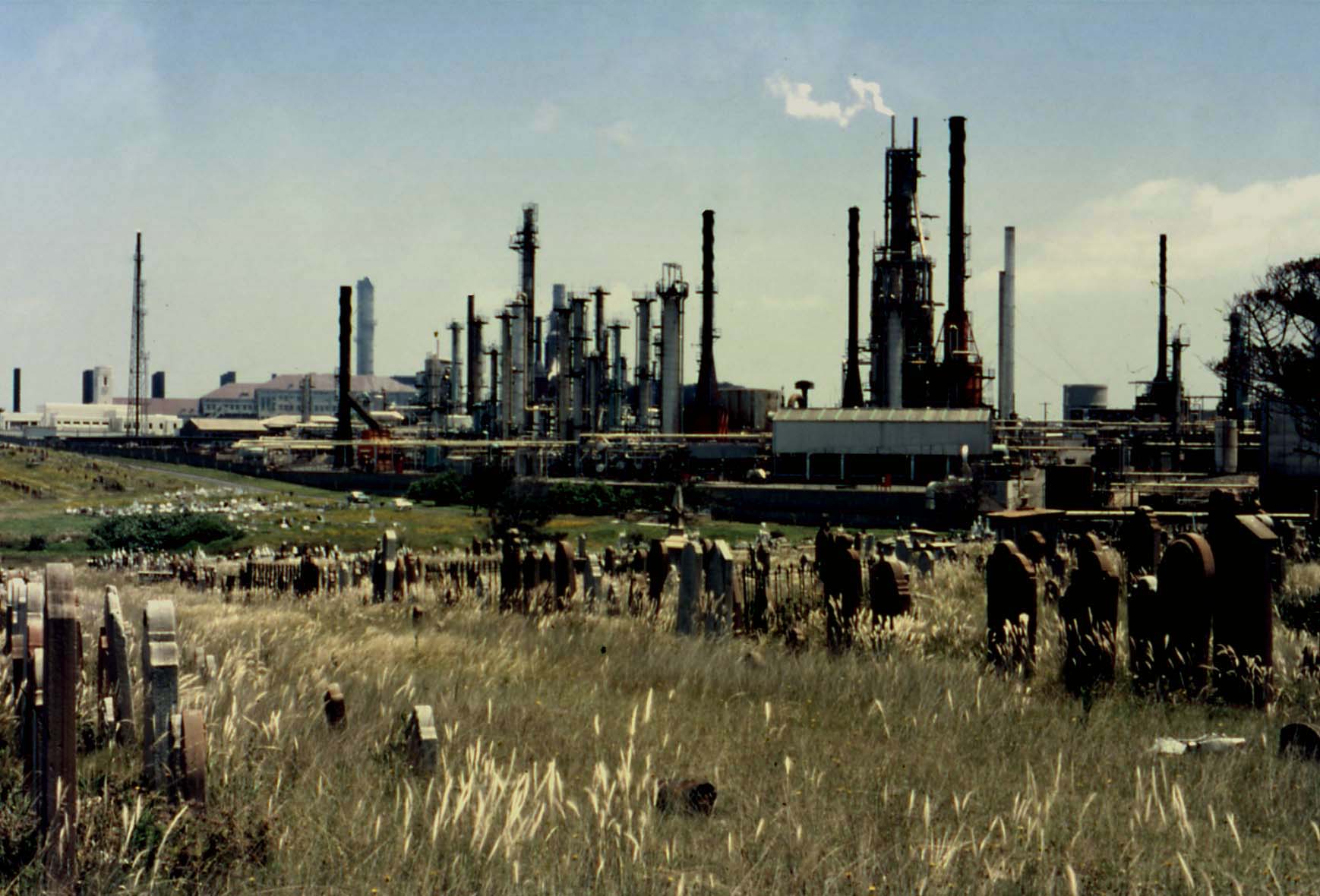 A cemetery filled with gravestones in the foreground, completely overshadowed by a massive, smoke-belching oil refinery looming immediately behind it.