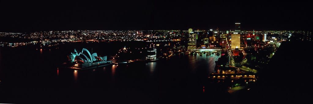 A panoramic night view of the Sydney Opera House glowing blue in the dark harbor, dwarfed by the illuminated towers of the financial district.