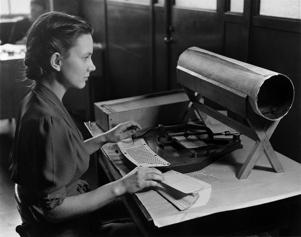 A woman operates a manual pantograph punch in a vintage photo, using a stylus to punch holes in a data card, illustrating the physical and industrial labor of early computing.