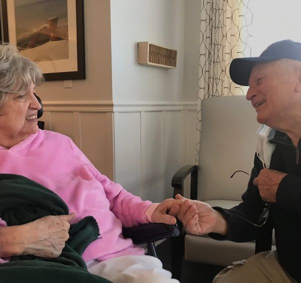Two elders with gray hair-- a woman in a pink shirt and a man in a blue coat and hat--hold hands and look lovingly at each other. 