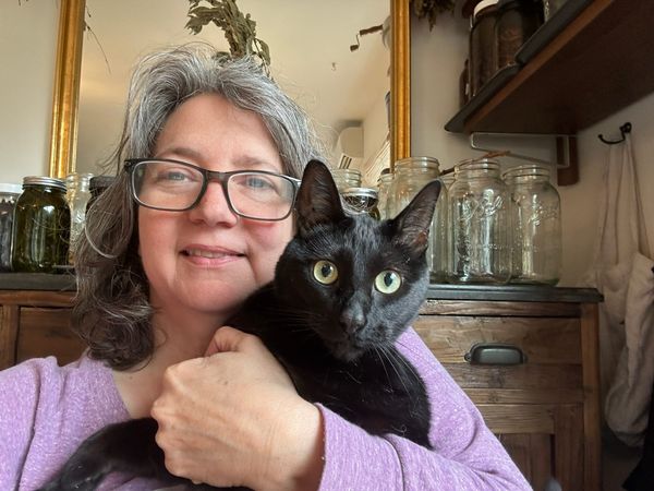 A photo of me--a fierce middle-aged white woman with glasses and gray hair--holding an even fiercer black cat starring straight at the camera 