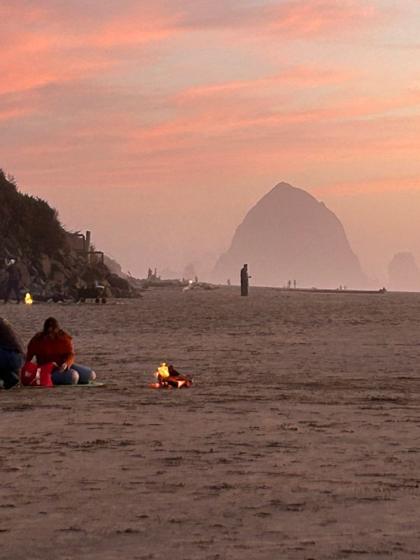 A photo of a beach at sunset with people in the distance sitting around fires and walking on the beach 