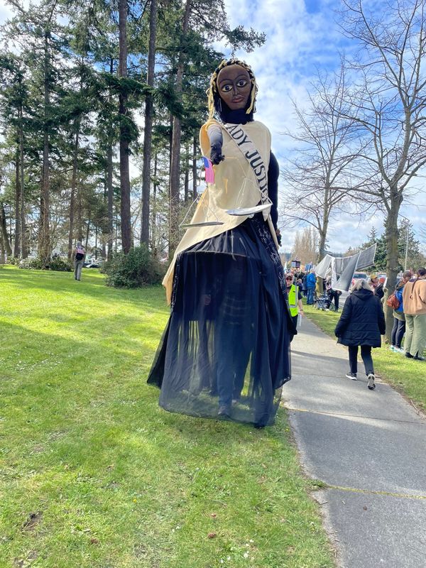 Photo of a 12-foot-tall Lady Justice costume at a No Kings protest. She's wearing a cream top with black skirt, holding the scales of justice, and has black skin and black and yellow hair.