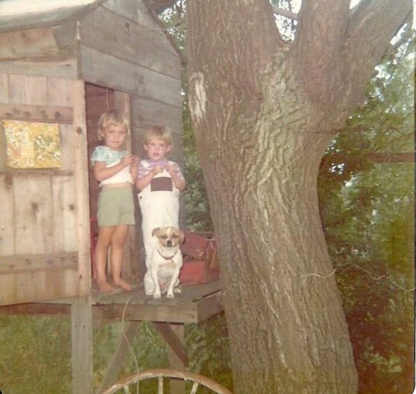 Old photo of two blond kids and a little white and brown dog standing in the door of an old unpainted treehouse 